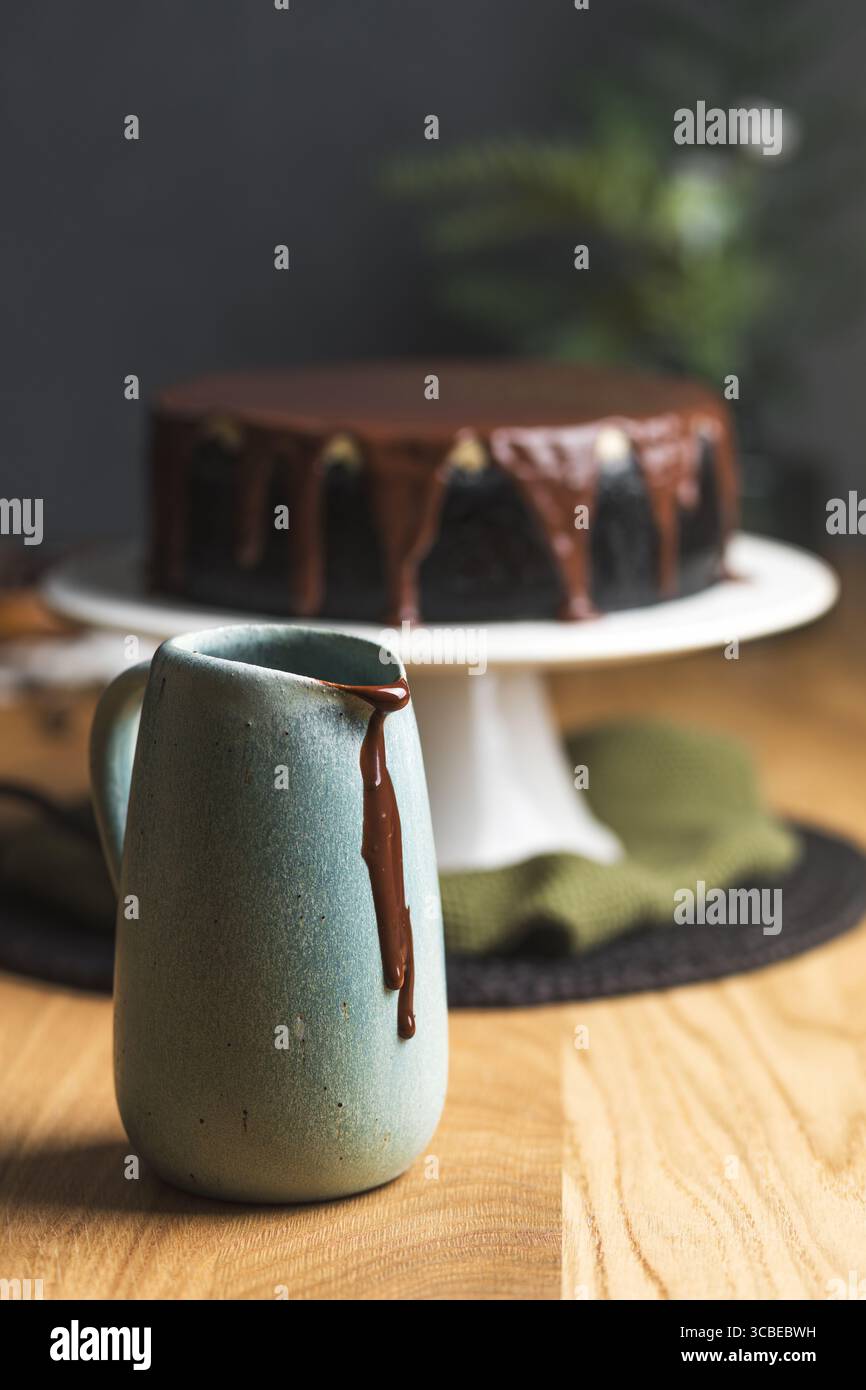 Una caraffa in ceramica con salsa di cioccolato che gocciola lateralmente, con una cheesecake ricoperta di ganache di cioccolato lucido sullo sfondo. Foto Stock