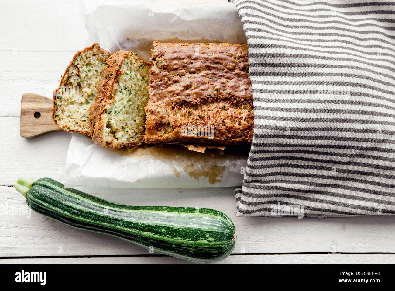 Ricetta per il pane di zucchine e formaggio, una pagnotta di pane su un tagliere di legno, ricoperta da un asciugamano da cucina, con accanto una zucchina intera. Foto Stock