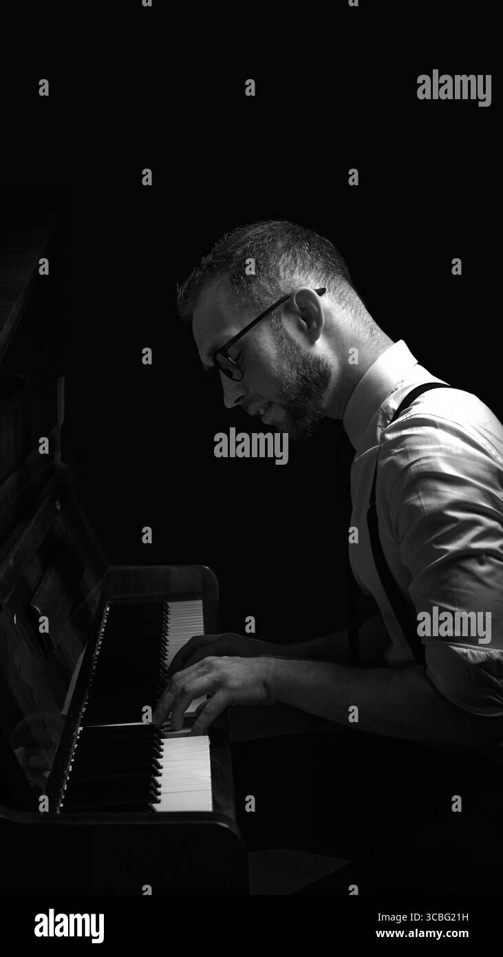 Uomo che suona il pianoforte con un'illuminazione soffusa e scura con occhiali e bretelle Foto Stock