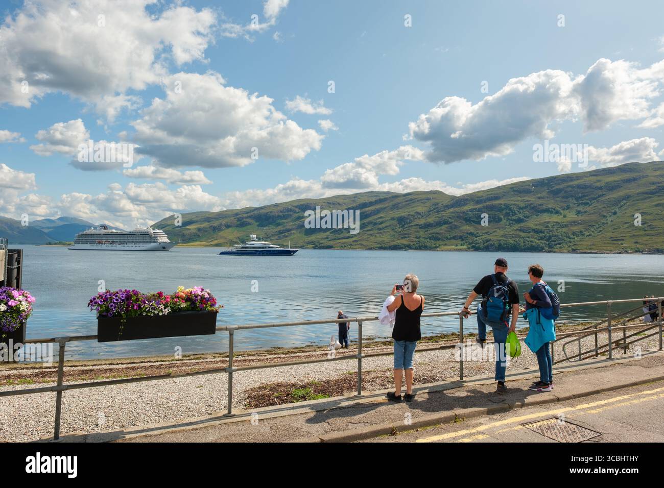 Persone sul lungomare di Ullapool, tra cui qualcuno che fotografa Loch Broom, con una nave da crociera in visita e uno yacht privato in viaggio contro le colline delle Highland. Foto Stock