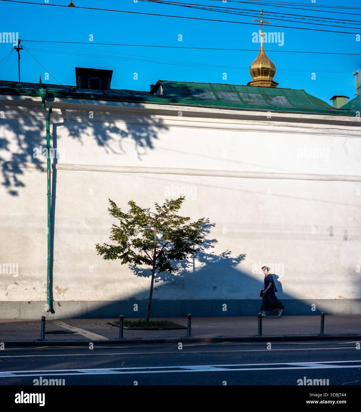 Una donna cammina su un marciapiede a Kiev vicino a una parete bianca di un edificio con una cupola dorata e un tetto verde, un piccolo albero sta gettando un'ombra Foto Stock
