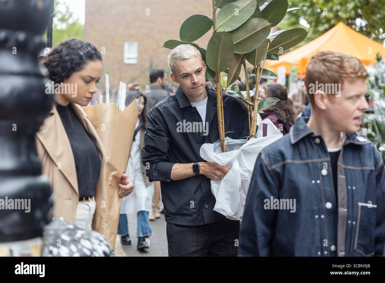 Londra, Regno Unito, 11 maggio 2025. Un uomo con capelli biondi sbiancati porta una pianta in vaso di Ficus elastica, Foto Stock