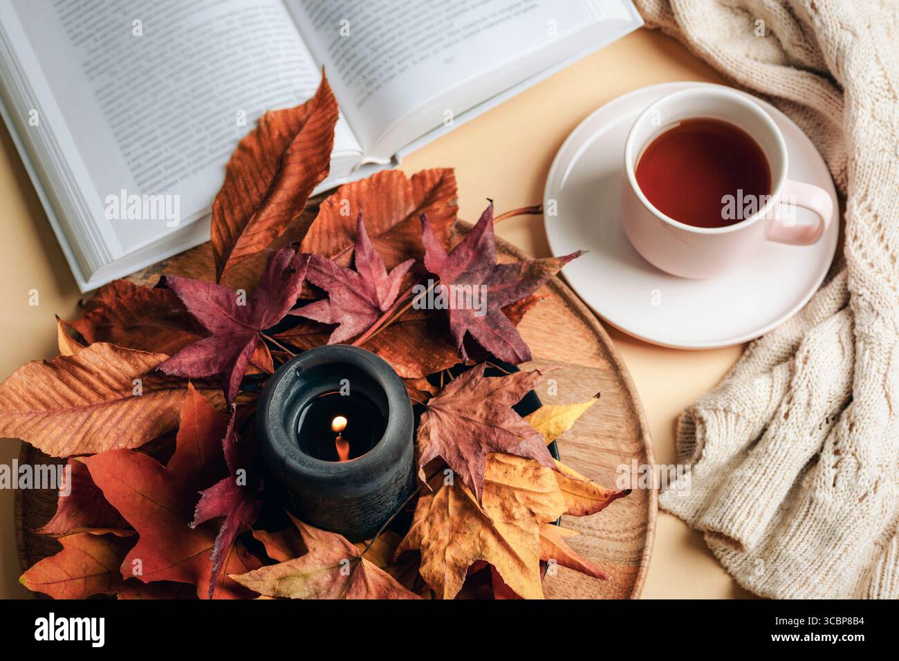 Tazza di tè con libro, candela nera e foglie autunnali su piatto di legno con maglione caldo. Atmosfera autunnale accogliente e relax. Vista dall'alto, disposizione piatta. Foto Stock