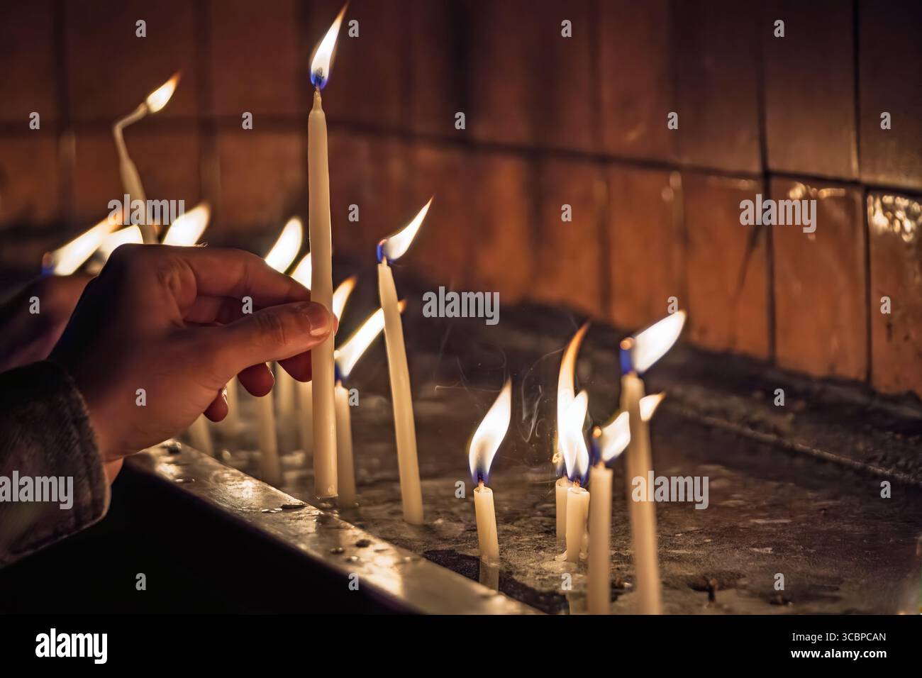 Un primo piano di una mano che posiziona con cura una candela accesa tremolante su un piedistallo, circondato da altre candele accese in un ambiente spirituale scarsamente illuminato Foto Stock