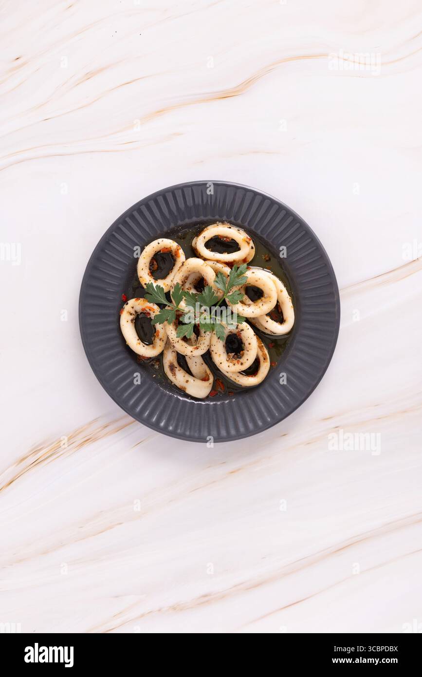 Anelli di calamaro decorati con erbe aromatiche, disposti in un motivo circolare su un piatto scuro su una superficie di marmo chiaro. M+S frutti di mare fotografati dall'alto. Foto Stock