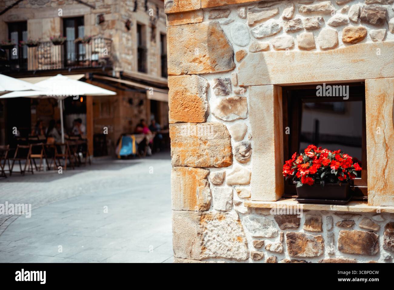 Rustica parete in pietra con vibranti fiori rossi in una finestra, affascinante Street Cafe con delicato sfondo focalizzato, calda luce del sole che crea un intimo Mediterraneo Foto Stock