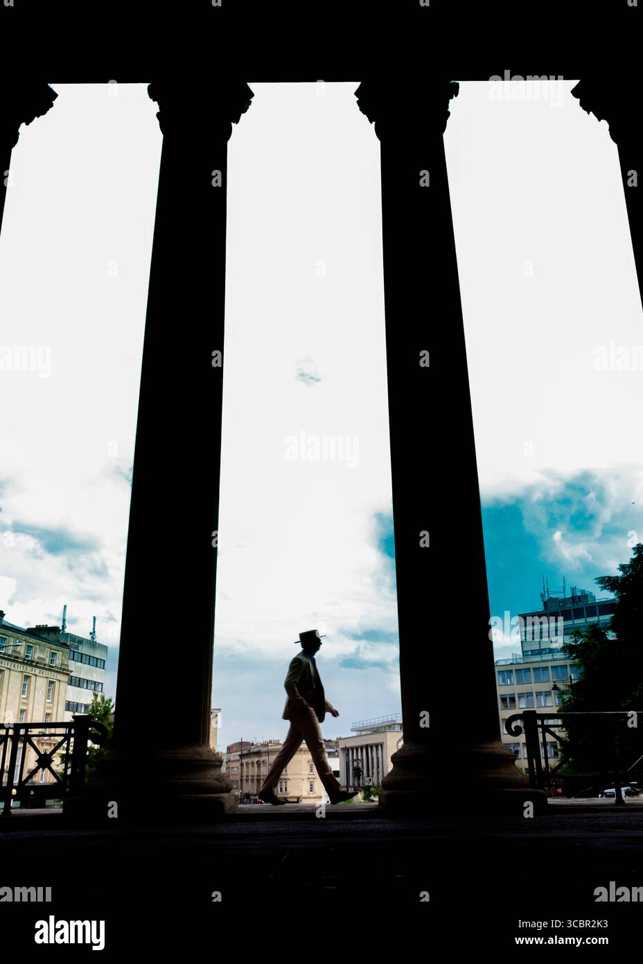 Un uomo sagomato con un abito e un cappello cammina tra grandi colonne. Foto Stock