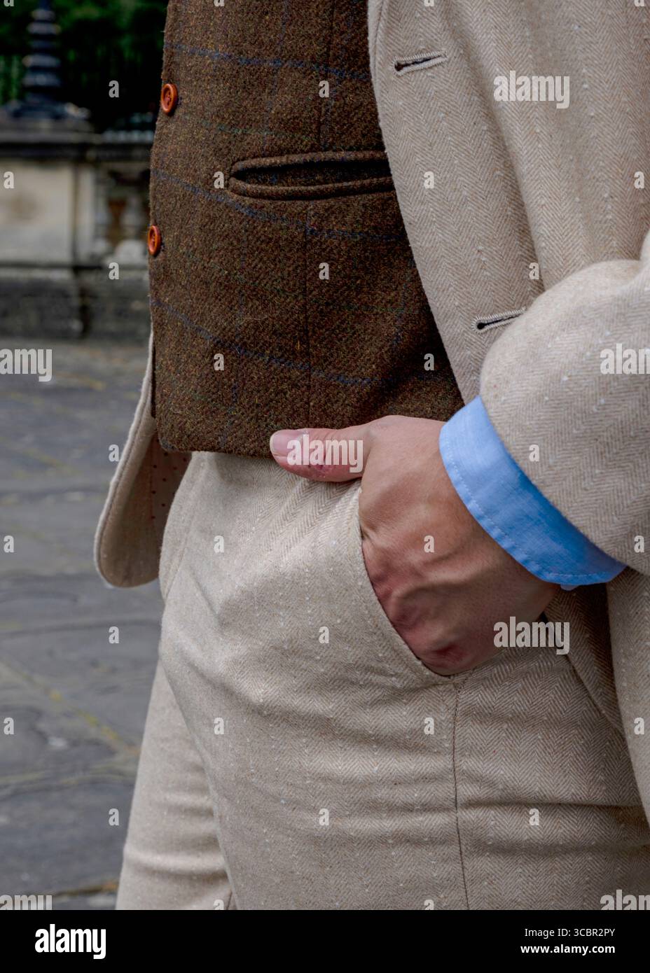 Un uomo che indossa un gilet marrone in tweed, una giacca da tuta di colore chiaro e pantaloni di colore chiaro, con la mano in tasca. Foto Stock