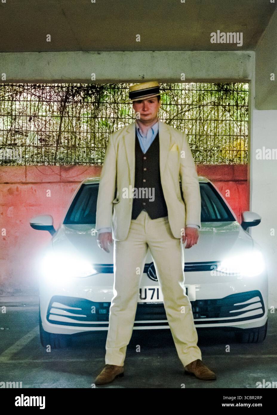 Un uomo con una tuta color crema e un cappello di paglia si trova di fronte a un'auto Volkswagen bianca con i fari accesi. Foto Stock