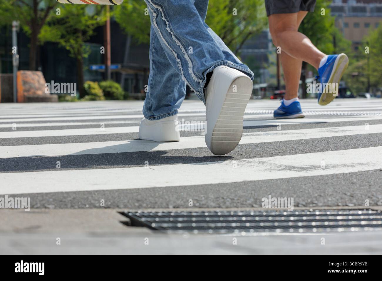 L'angolo basso di due persone che attraversano l'intersezione mostra il movimento. Caratterizzato da una marcia media, mette in risalto gli outfit casual urbani: Jeans blu e abbigliamento da running. Foto Stock