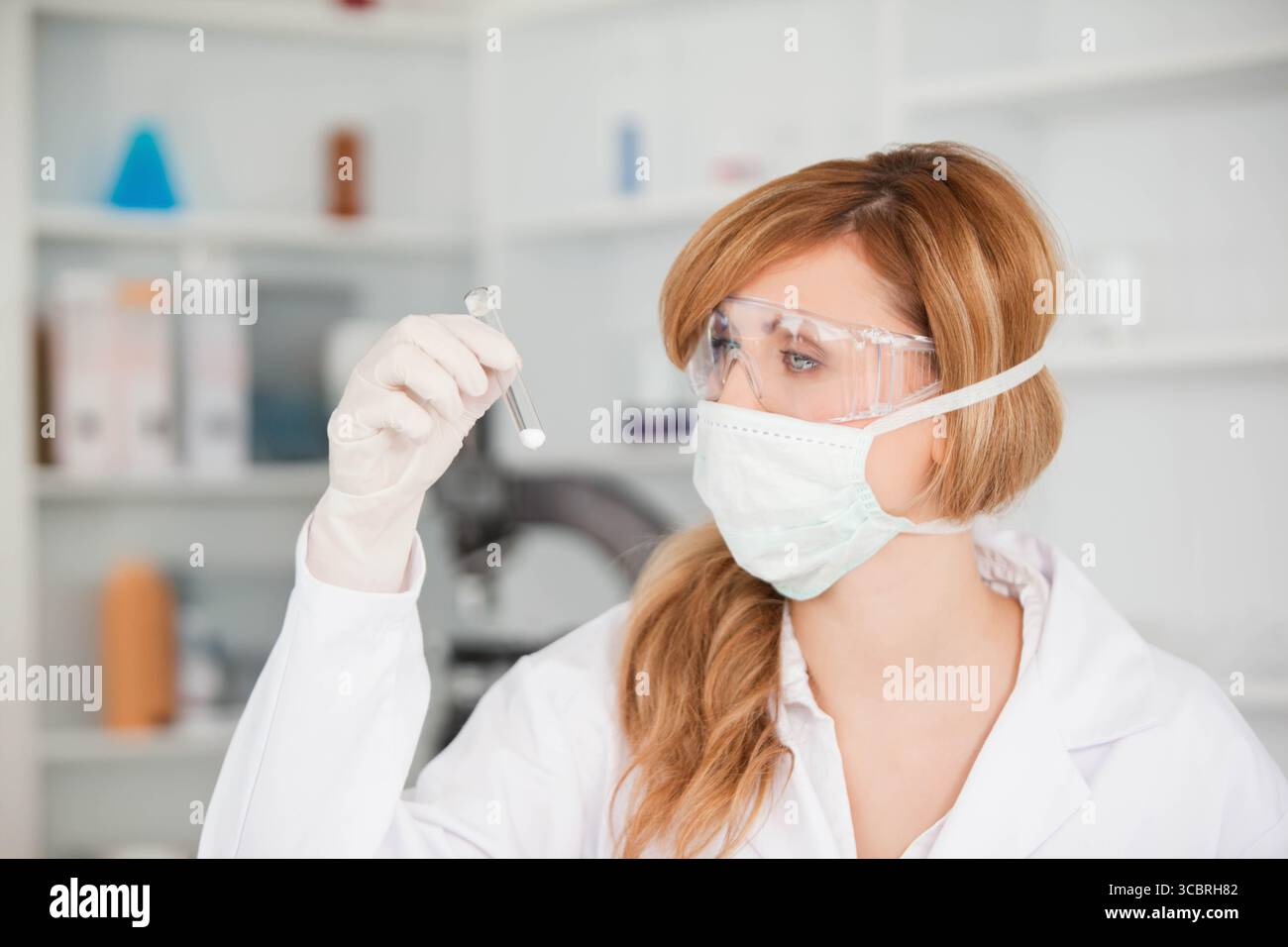 Ricercatrice di laboratorio che indossa dispositivi di protezione per l'esame della provetta in laboratorio sterile con microscopio Foto Stock