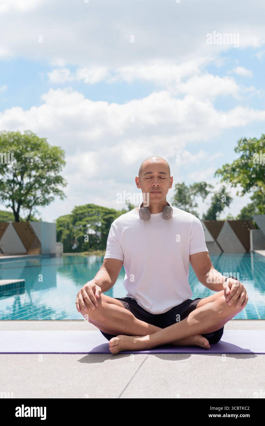 L'uomo calvo latino seduto in meditazione posa a bordo piscina. Momento di calma e tranquillità che riflette yoga, consapevolezza, concentrazione spirituale e sana vita estiva Foto Stock
