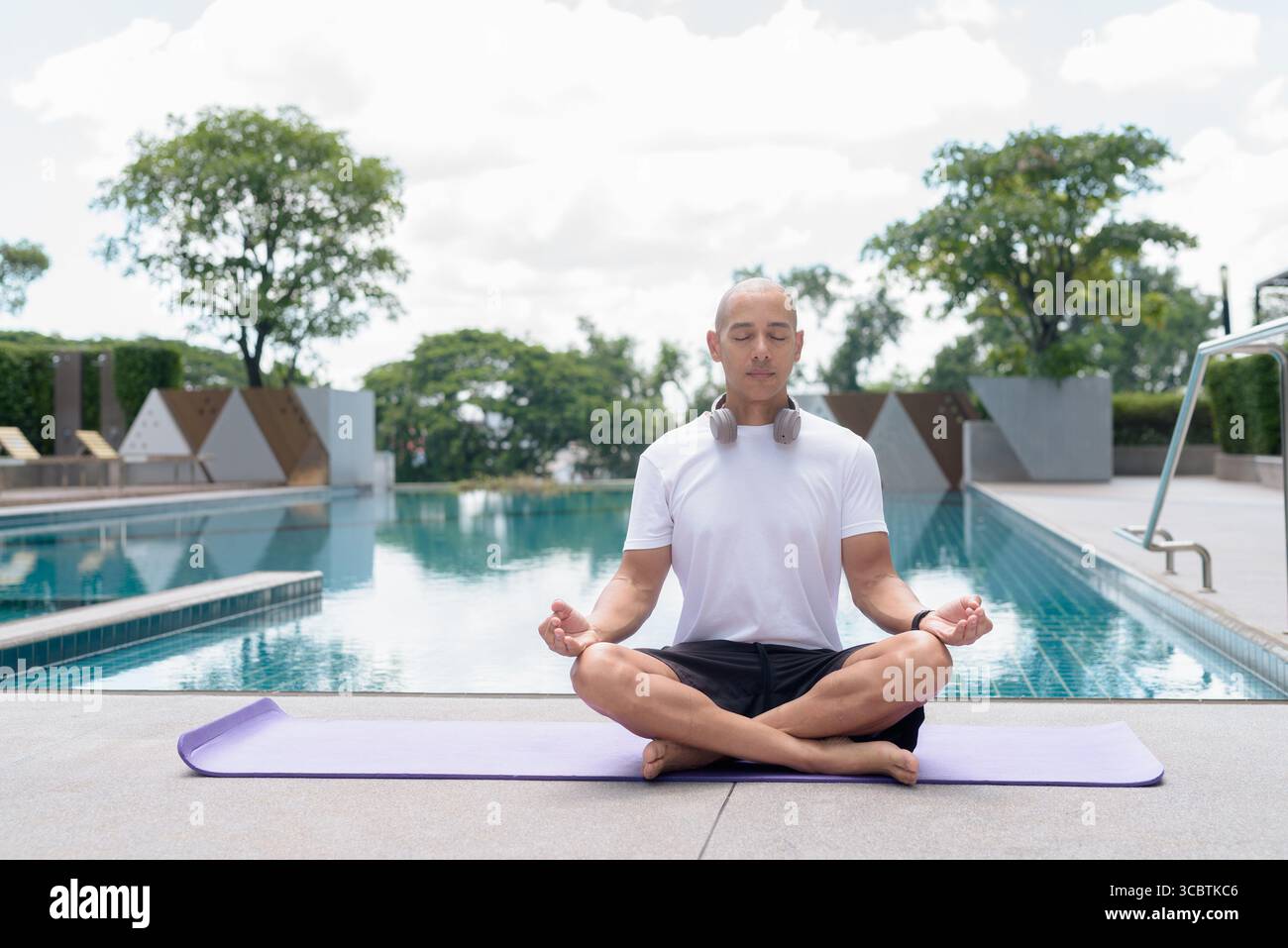 L'uomo calvo latino seduto in meditazione posa a bordo piscina. Momento di calma e tranquillità che riflette yoga, consapevolezza, concentrazione spirituale e sana vita estiva Foto Stock