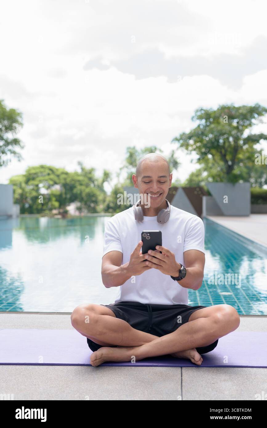 Uomo calvo latino seduto in meditazione posare a bordo piscina mentre usa lo smartphone. Momento di calma e tranquillità che riflette yoga, consapevolezza, concentrazione spirituale a. Foto Stock