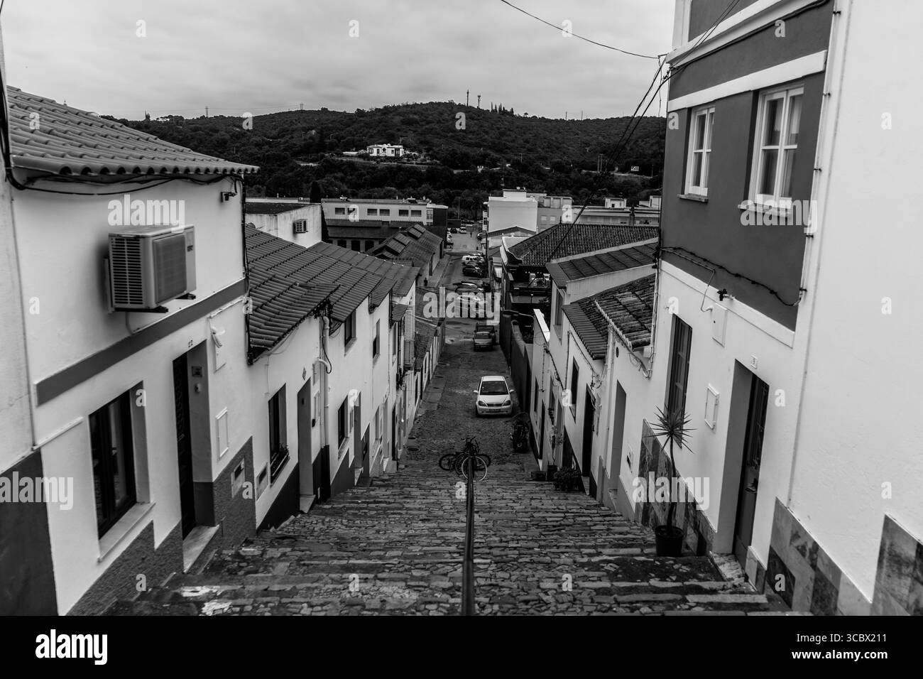 Algarve, Portogallo – 2025.07.04: Vista da una ripida scalinata acciottolata fiancheggiata da case colorate e tetti di tegole rosse, che conduce a una strada con parke Foto Stock