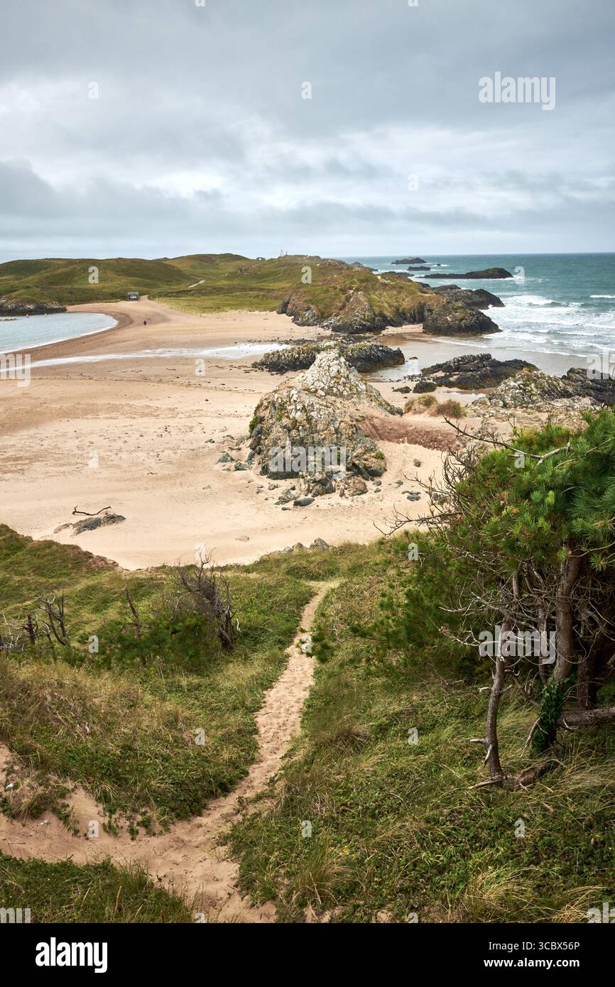 Il mare taglia al largo dell'isola di Llanddwyn dalla terraferma di Anglesey su una stretta striscia di sabbia con flussi di lava a cuscino in primo piano. Foto Stock