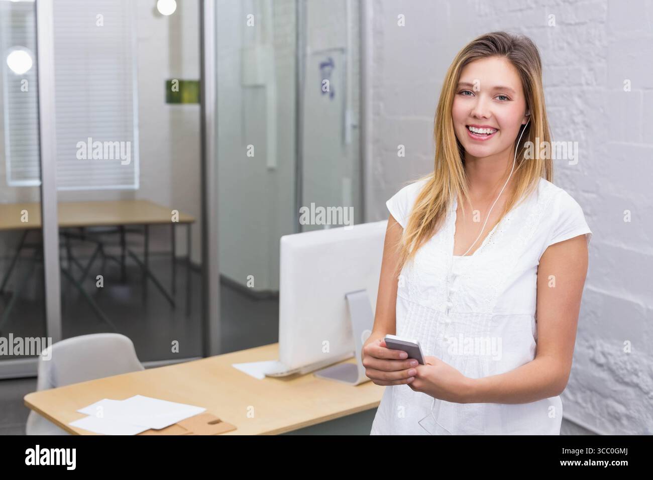 Dipendente di un ufficio in piedi accanto alla scrivania con monitor che tiene lo smartphone e gli auricolari spazio di copia Foto Stock
