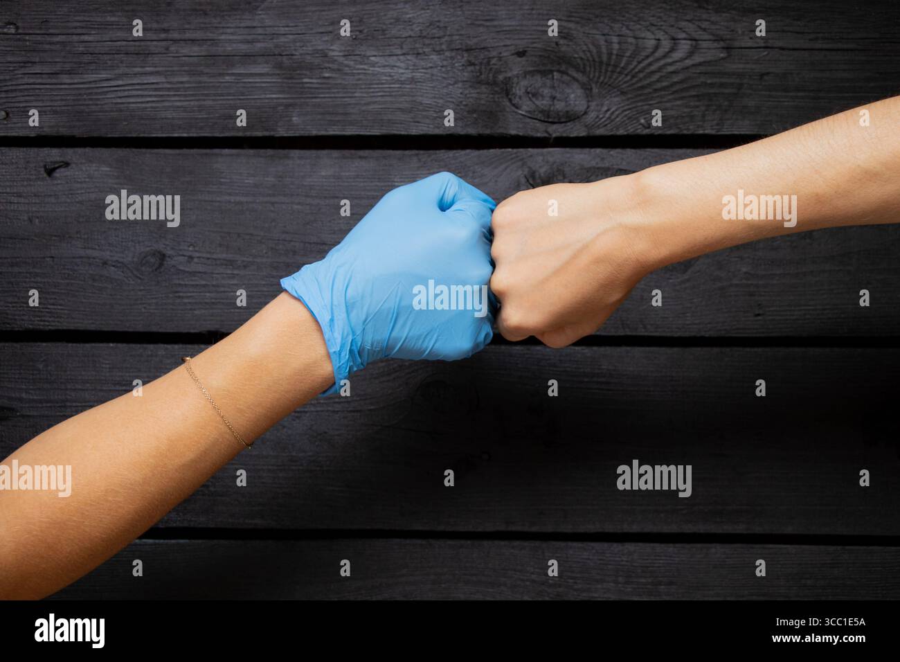 Una mano in un guanto medico blu e una mano femminile senza guanto bump pugni su uno sfondo di legno nero Foto Stock