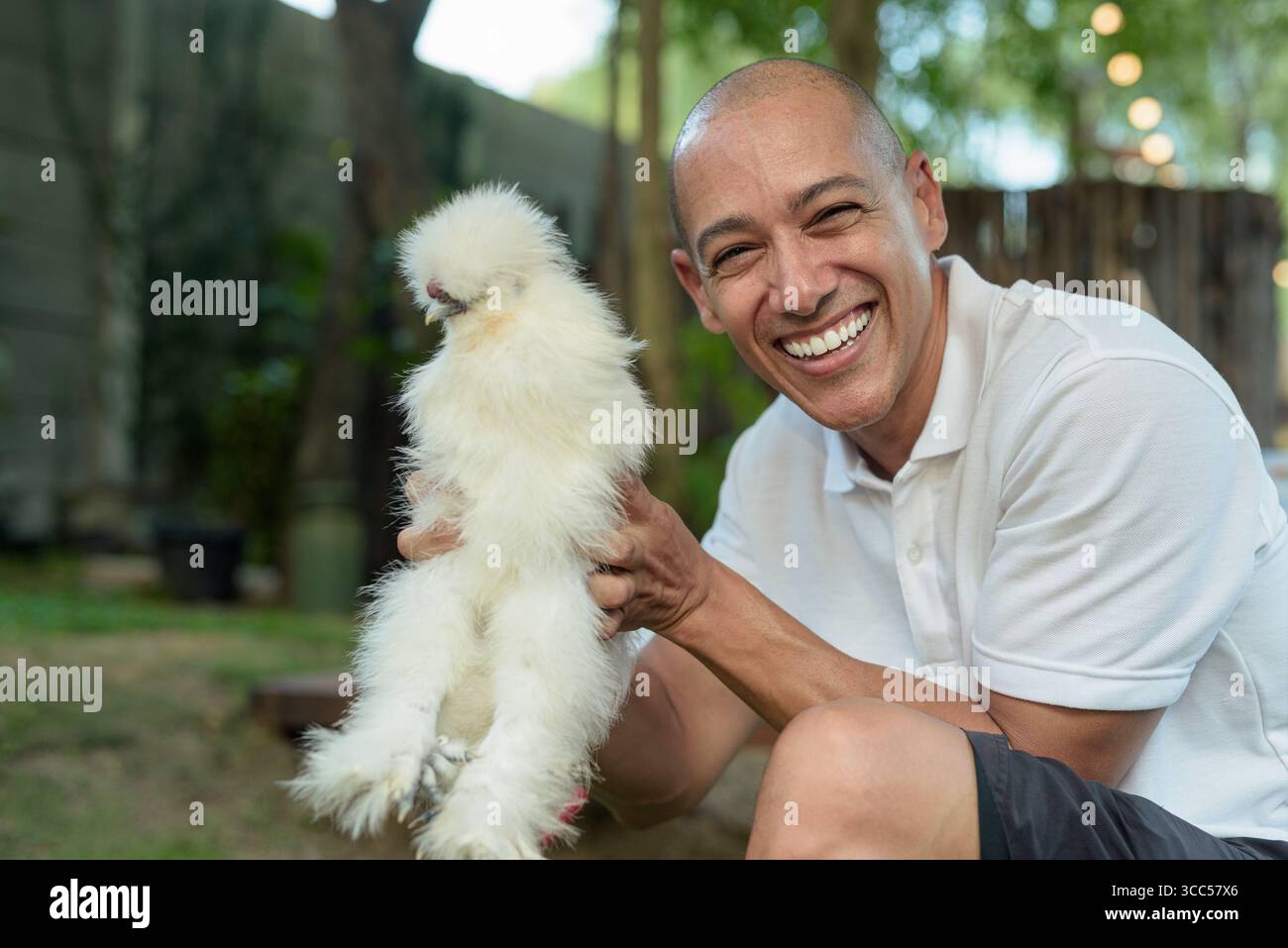 Uomo calvo con una polo bianca sorridente calorosamente mentre tiene in mano un amico di pollo all'aperto. Ritratto concettuale che esprime gentilezza, compassione animale, e. Foto Stock