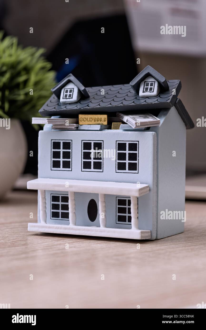 Modello di piccola casa con tetto sollevato per rivelare fasci di contanti e lingotti d'oro, simboleggiando gli investimenti immobiliari e la ricchezza nascosta. Foto Stock