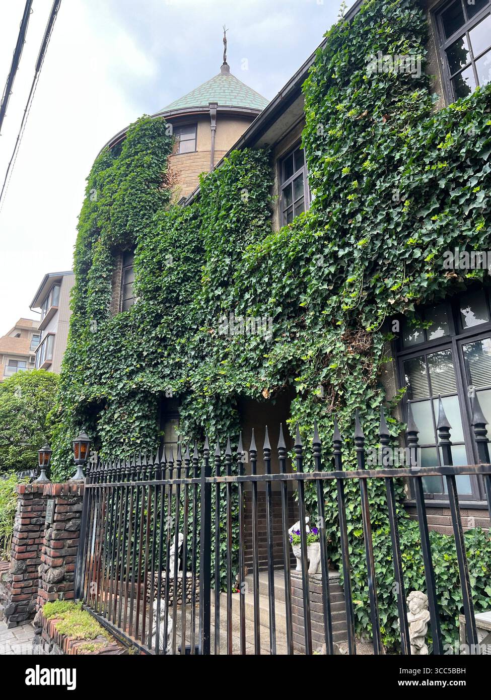 Residenza storica rivestita di edera con torretta e recinzione in ferro battuto su una strada urbana, mattoni e stucchi avvolti nel verde. Foto Stock