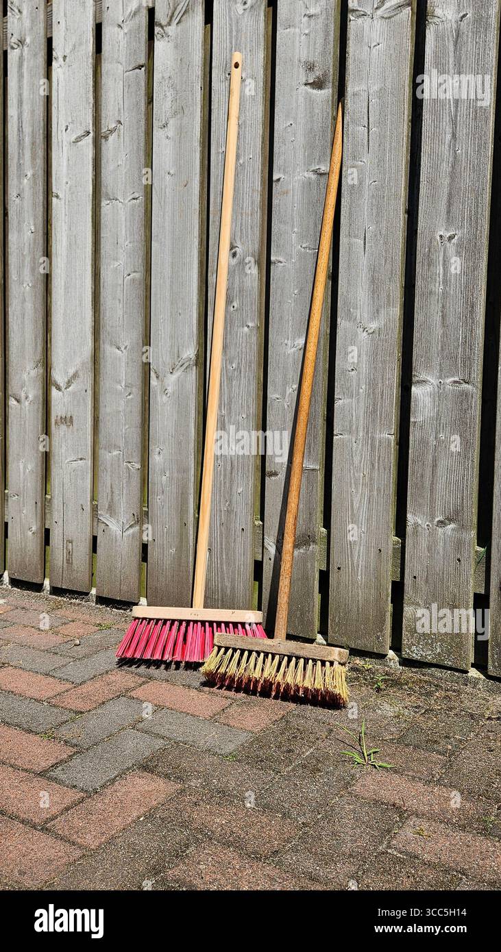 Due scope con manici in legno e setole colorate appoggiate su una recinzione rustica in legno su un patio in mattoni. - Immagine stock catturata con smartphone