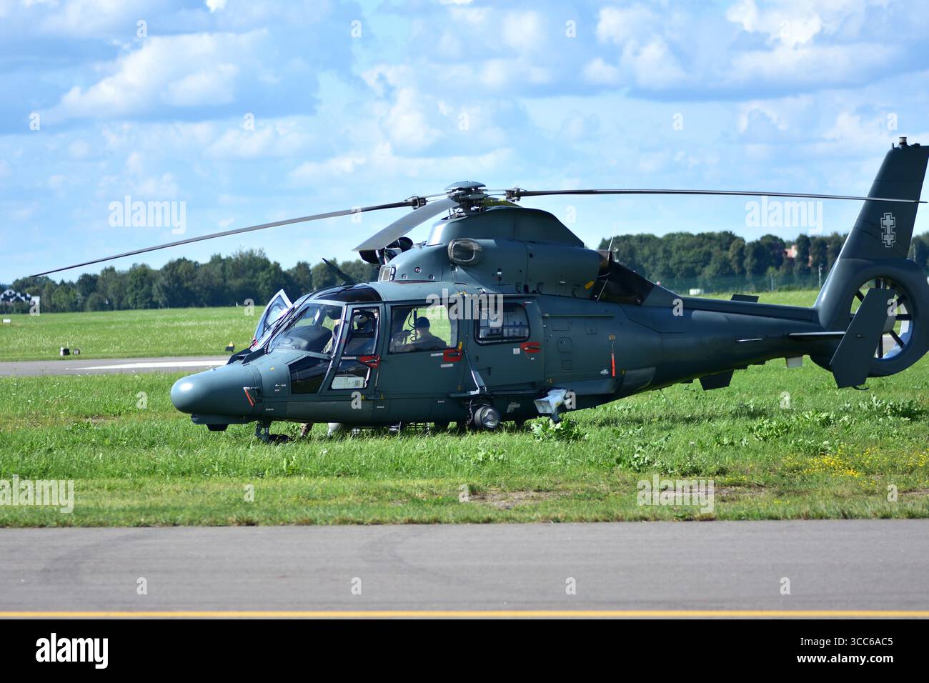 Elicottero militare verde scuro parcheggiato su un campo d'aviazione erboso, vista laterale con porta della cabina di pilotaggio aperta, piloti visibili all'interno, sotto un cielo azzurro luminoso con scattere Foto Stock