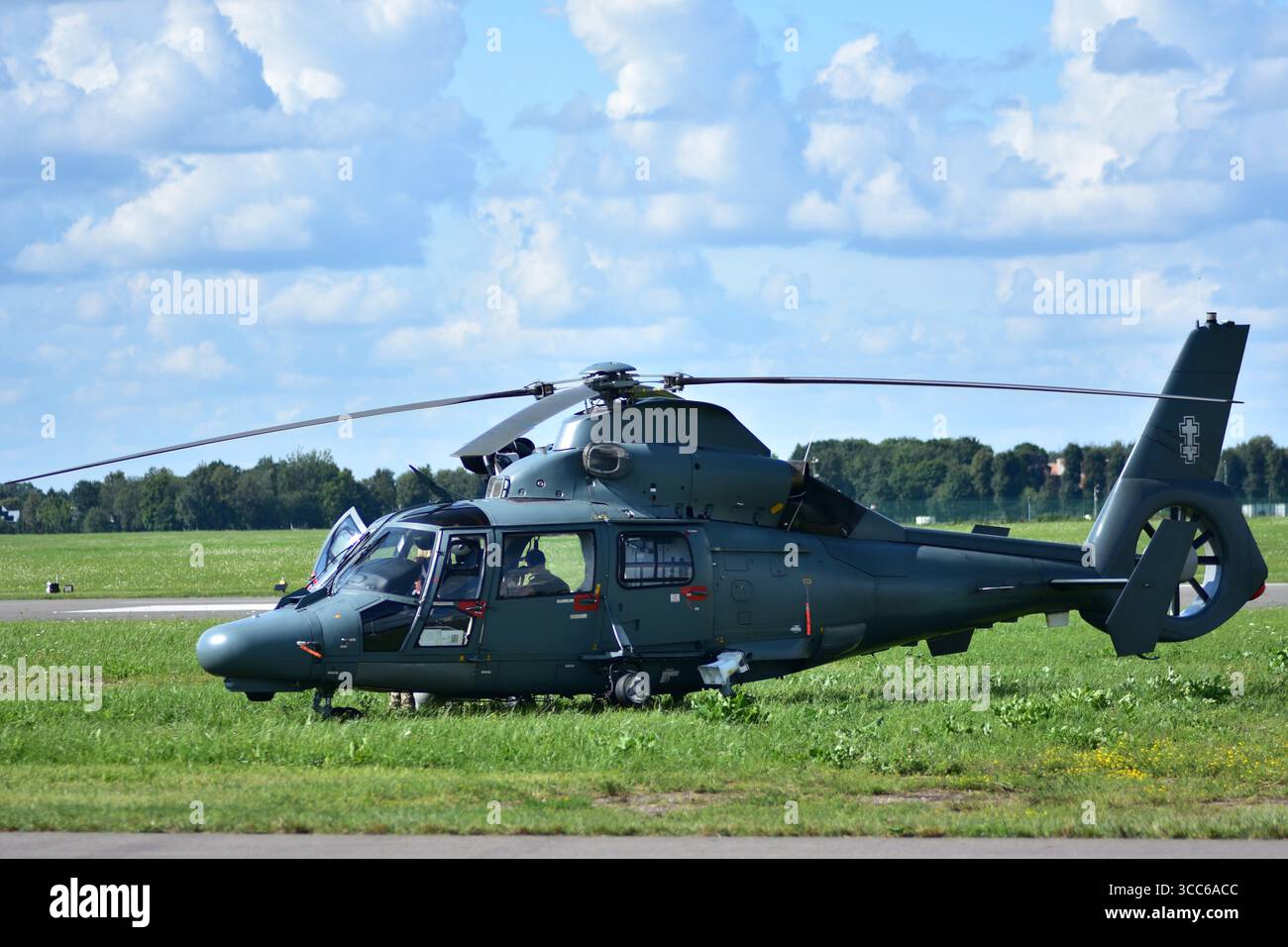 Elicottero militare verde scuro parcheggiato su un campo d'aviazione erboso, vista laterale con porta della cabina di pilotaggio aperta, piloti visibili all'interno, sotto un cielo azzurro luminoso con scattere Foto Stock