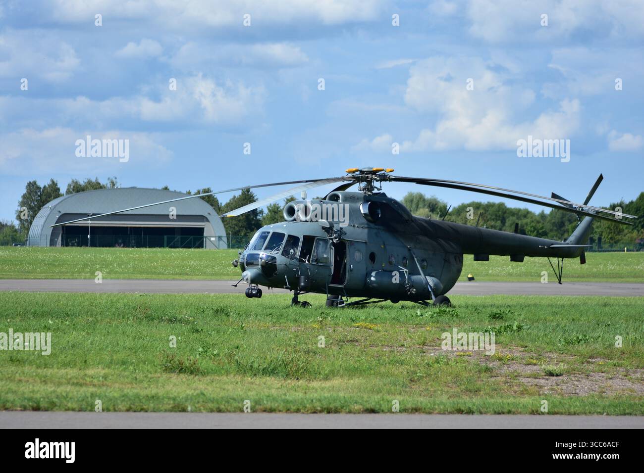 Grande elicottero da trasporto militare mi-17 su un campo d'aviazione erboso, vista frontale con pale del rotore estese, finestre del cockpit che riflettono la luce del sole, hangar in ba Foto Stock