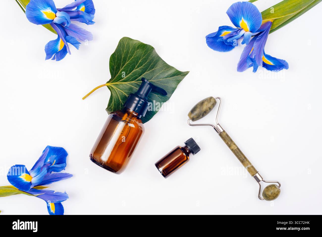 Bottiglie cosmetiche marroni, rullo facciale, fiore di Iris blu e foglia verde su sfondo bianco. Vista dall'alto, disposizione piatta. Foto Stock
