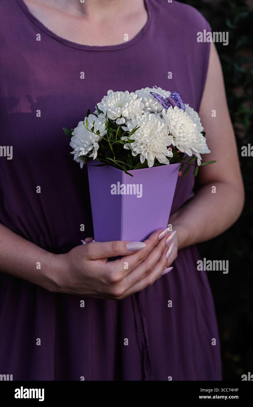 Elegante bouquet di crisantemo bianco in scatola regalo viola con vegetazione lussureggiante e farfalla ornamentale. Decorazioni floreali moderne per saluti, feste o feste Foto Stock