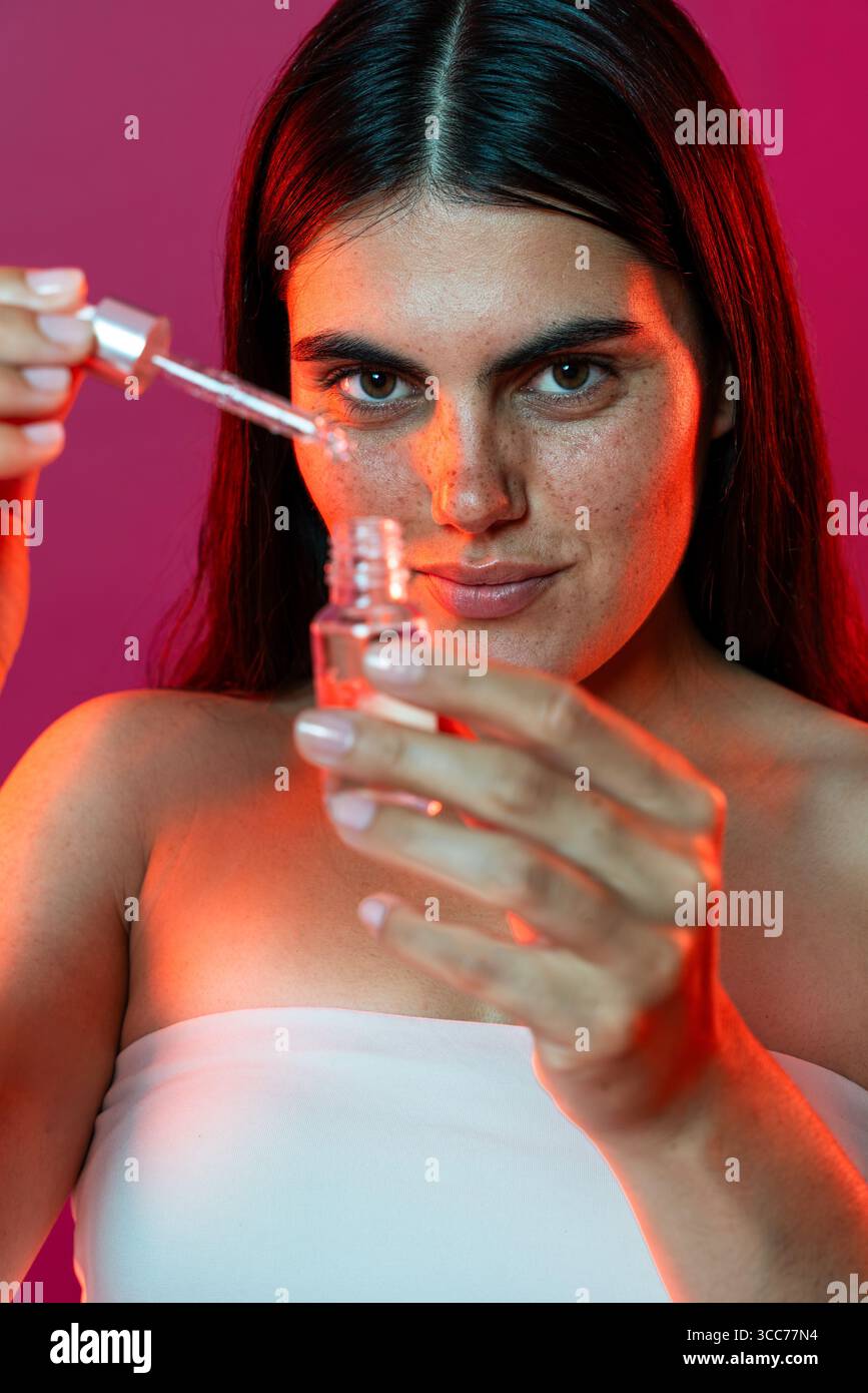 Una giovane donna con lentiggini tiene in mano una bottiglia di vetro e un contagocce, applicando con cura il siero facciale come parte della sua routine di cura della pelle in un vivace ambiente da studio Foto Stock