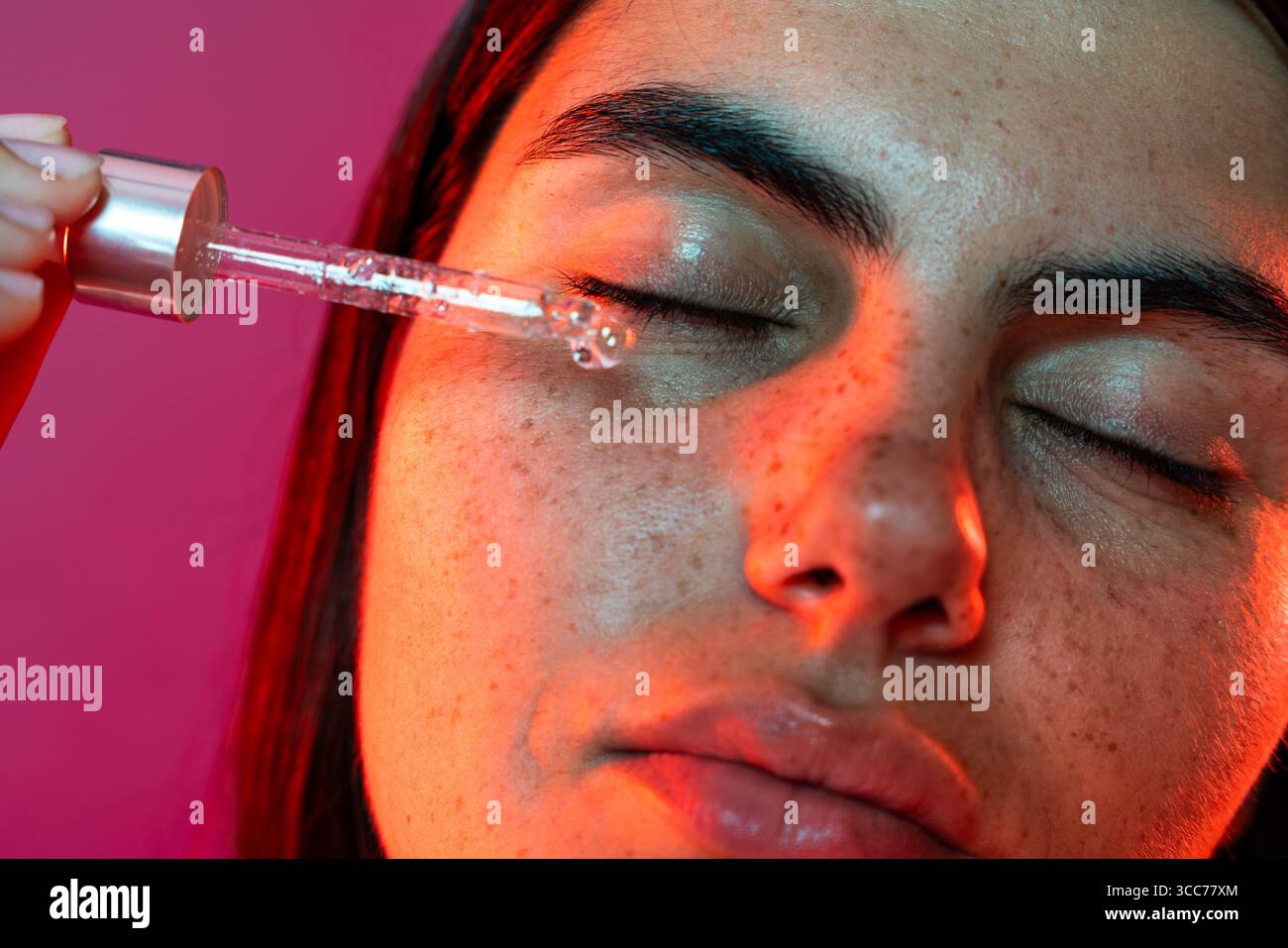 Giovane donna con lentiggini che si gode un trattamento facciale, applicando siero con un contagocce e chiudendo gli occhi in uno studio rosa illuminato da soffici lori rossi Foto Stock