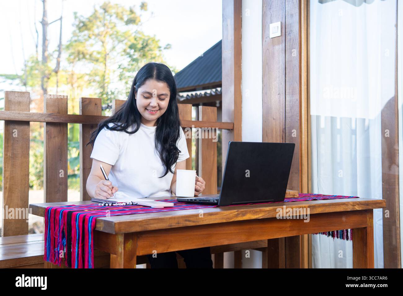 Una giovane donna asiatica con una t-shirt bianca lavora su un computer portatile presso una scrivania di legno all'aperto, scrivendo appunti e sorridendo. Lavoro in remoto, stile di vita freelance. Foto Stock