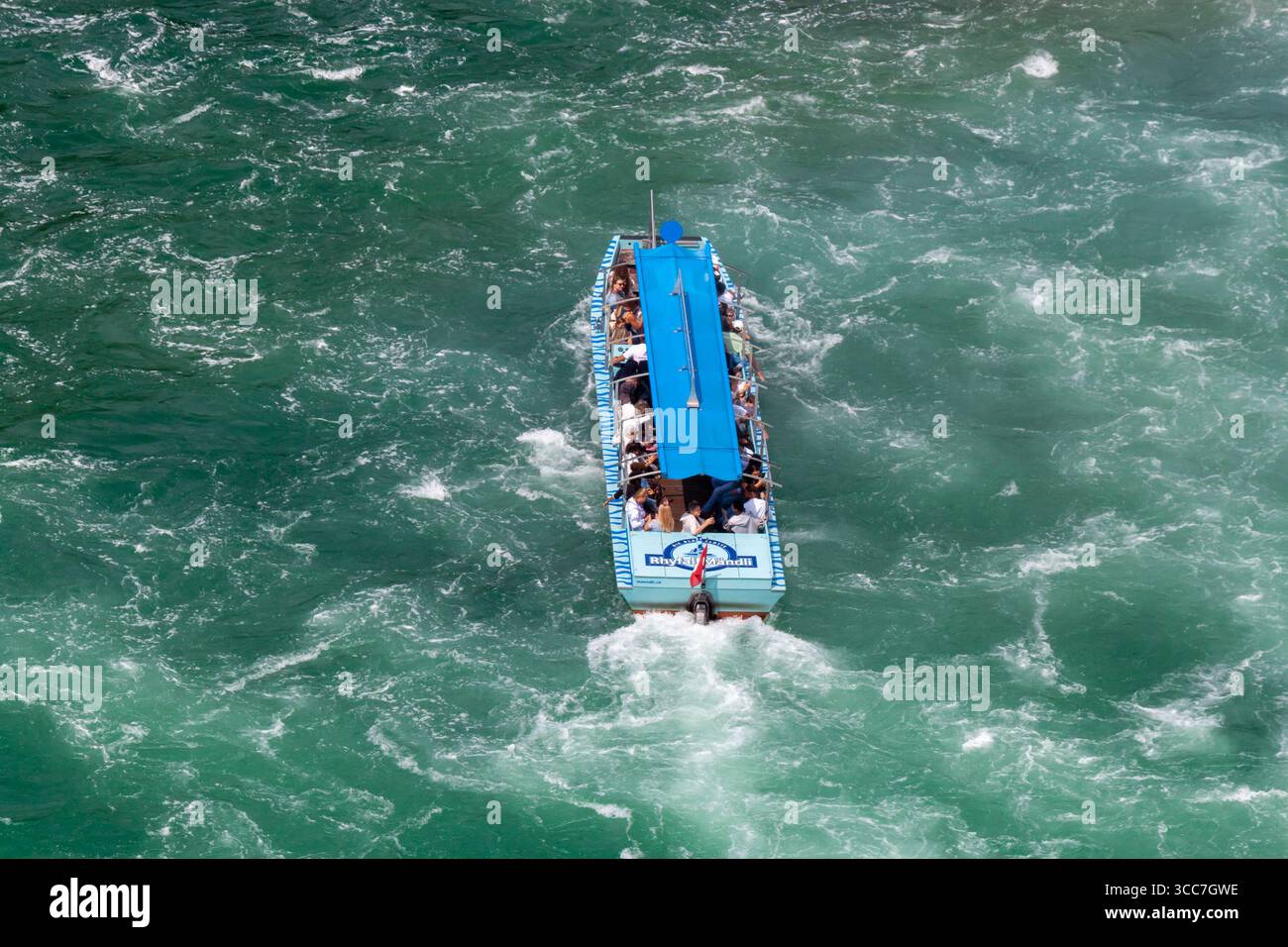 NEUHAUSEN AM RHEINFALL, SVIZZERA - 6 AGOSTO 2025: Una piccola barca turistica blu, piena di gente, naviga nelle turbolente acque turchesi della R. Foto Stock