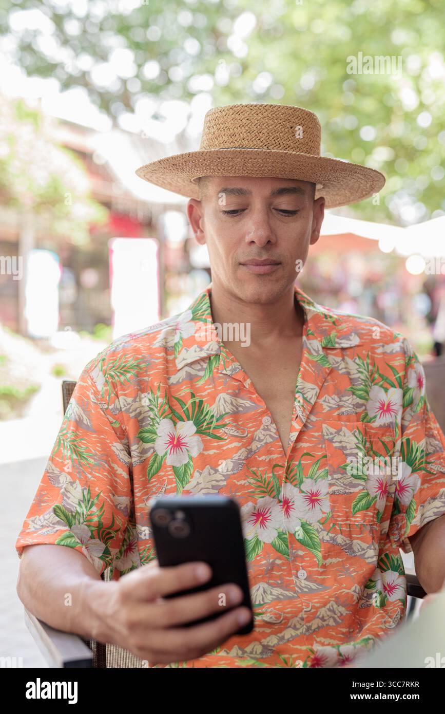Un turista ispanico di sesso maschile seduto a un tavolino all'aperto in una giornata di sole, indossando un cappello Panama e una camicia a manica corta mentre utilizza il suo smartphone. Casua Foto Stock