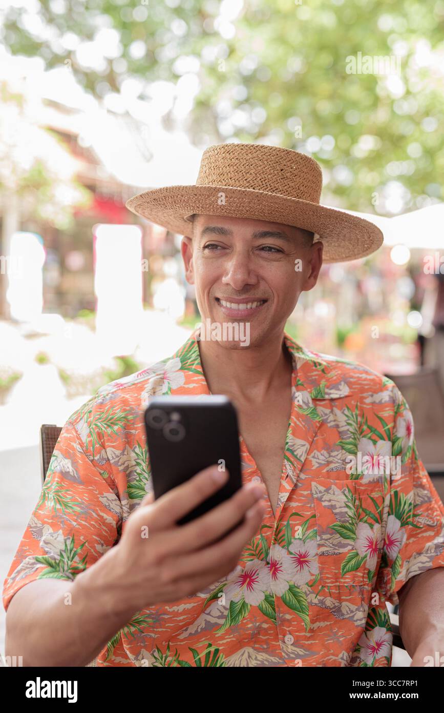 Un turista ispanico di sesso maschile seduto a un tavolino all'aperto in una giornata di sole, indossando un cappello Panama e una camicia a manica corta mentre utilizza il suo smartphone. Casua Foto Stock