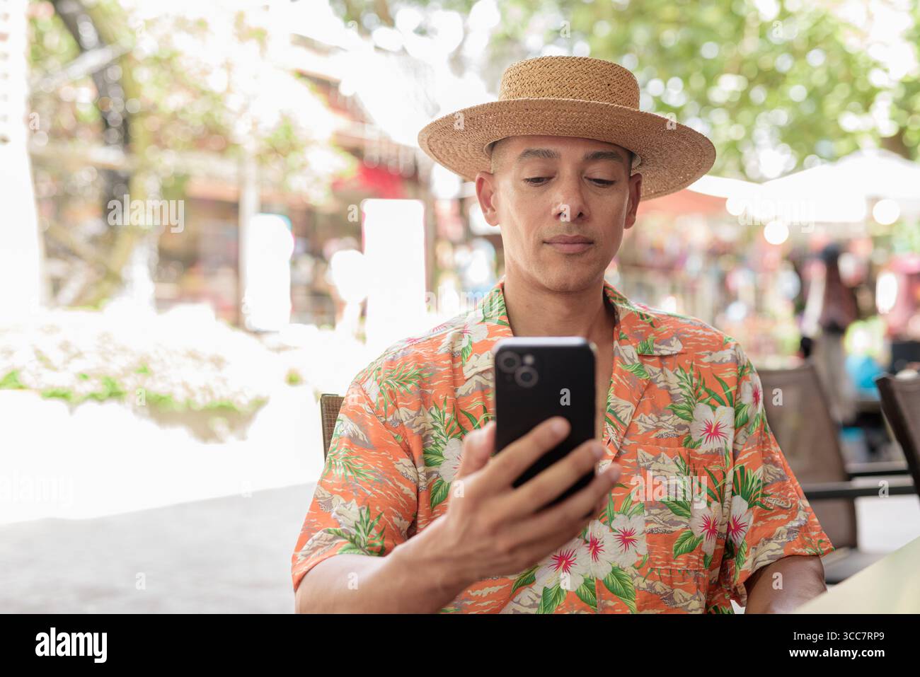 Un turista ispanico di sesso maschile seduto a un tavolino all'aperto in una giornata di sole, indossando un cappello Panama e una camicia a manica corta mentre utilizza il suo smartphone. Casua Foto Stock