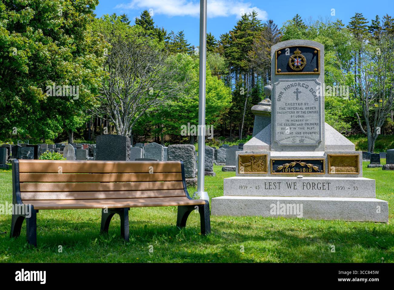 Saint John, NB, Canada - 21 giugno 2025: Un monumento di guerra canadese dice "OUR HONED DEAD" e "AT WE FORGET" in un cimitero. Una panchina del parco è accanto al Th Foto Stock