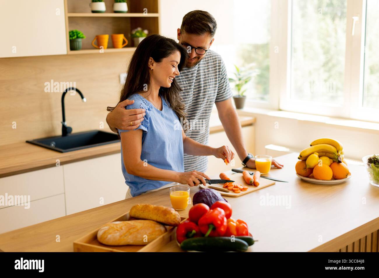 Una coppia gioiosa condivide un momento speciale nella loro cucina luminosa, tagliando verdure e godendosi la compagnia dell'altra mentre preparano un pasto sano Foto Stock