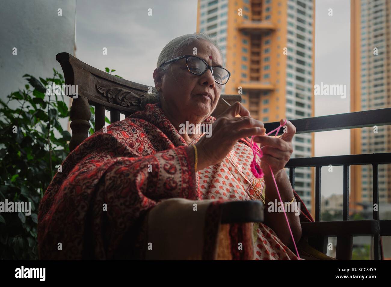 Anziana donna indiana seduta su una sedia a dondolo in legno su un balcone, maglieria con palline colorate in filato, con un tradizionale sari e occhiali. Foto Stock