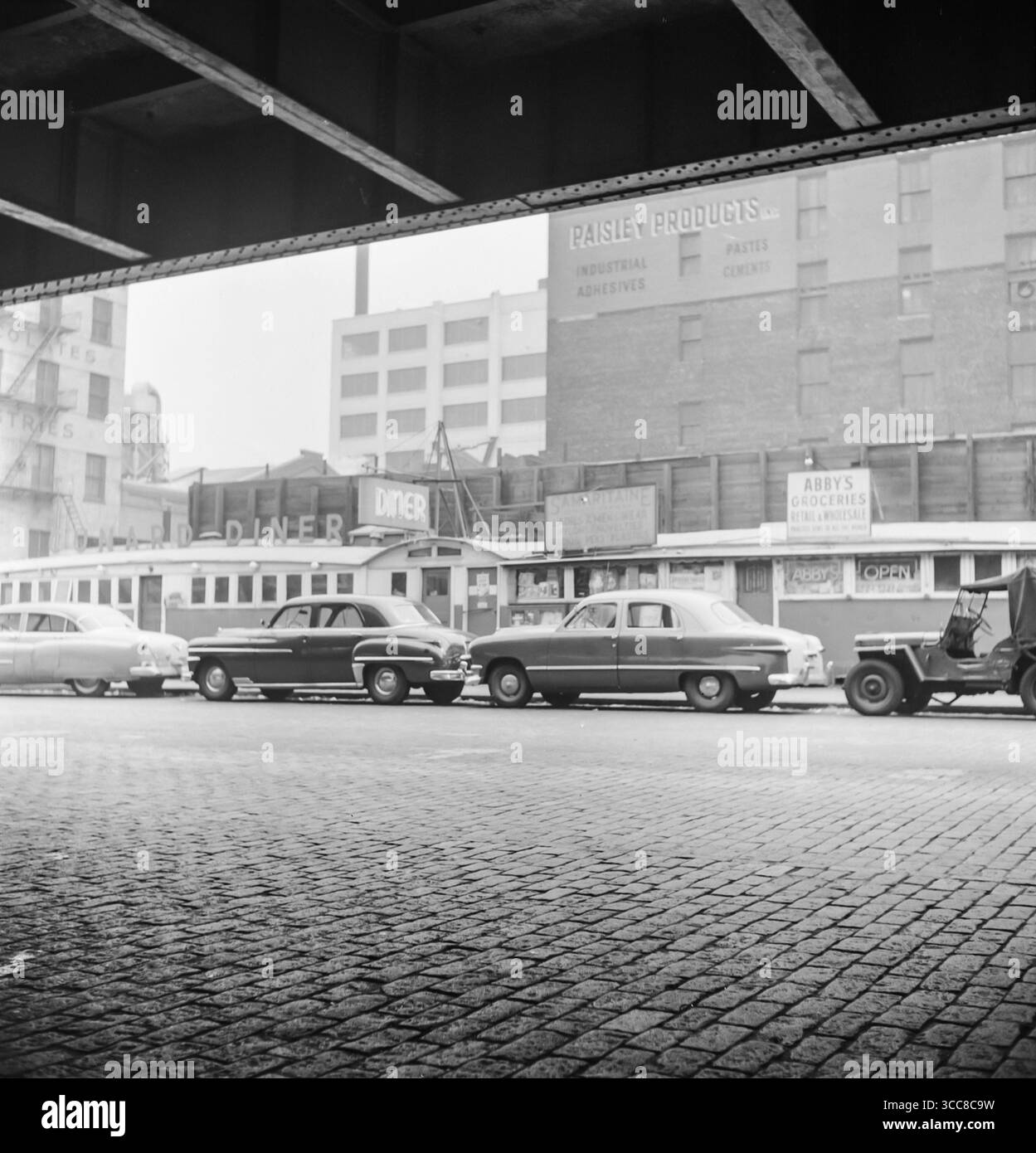 Una fotografia vintage in bianco e nero del 1952 che mostra una fila di edifici vicino al porto di New York. Ci sono i negozi di alimentari Cunard Diner e Abby, e aline di auto parcheggiate. Foto Stock