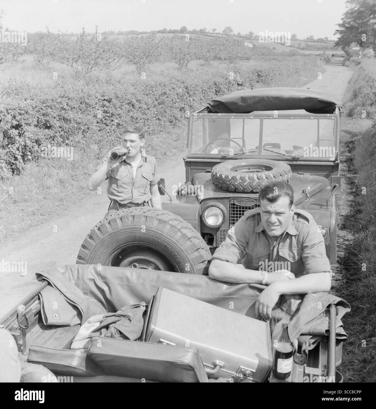 Verso la fine degli anni '1950 fotografia in bianco e nero che mostra due membri maschi dell'esercito britannico di fronte a una Land Rover serie 1, su una strada di campagna in Inghilterra. Un uomo sta bevendo una bottiglia di birra. Foto Stock