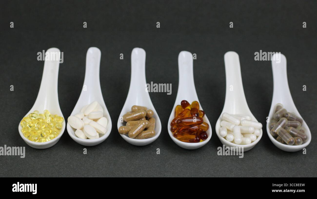 Sei cucchiai bianchi ripieni di integratori assortiti, vitamine e capsule visualizzati in una riga ordinata su sfondo grigio. Foto Stock