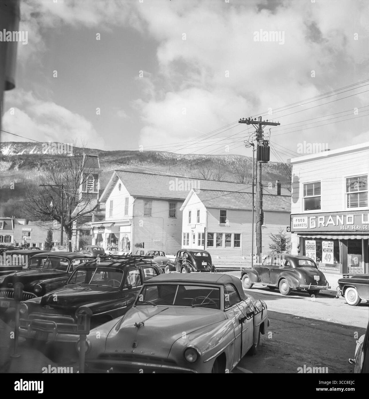 Una fotografia vintage in bianco e nero del 1952 che mostra una città in America con auto lungo le strade e le colline sullo sfondo e negozi lungo le strade. Foto Stock