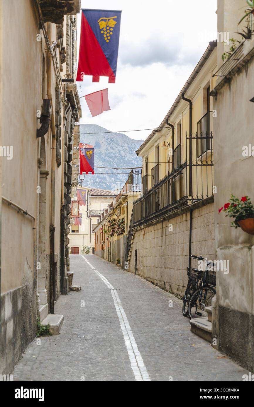 Veduta della stretta strada fiancheggiata da vecchi edifici e striscioni, che porta in lontananza, con montagne che si affacciano oltre Sulmona, Abruzzo, Italia. Foto Stock
