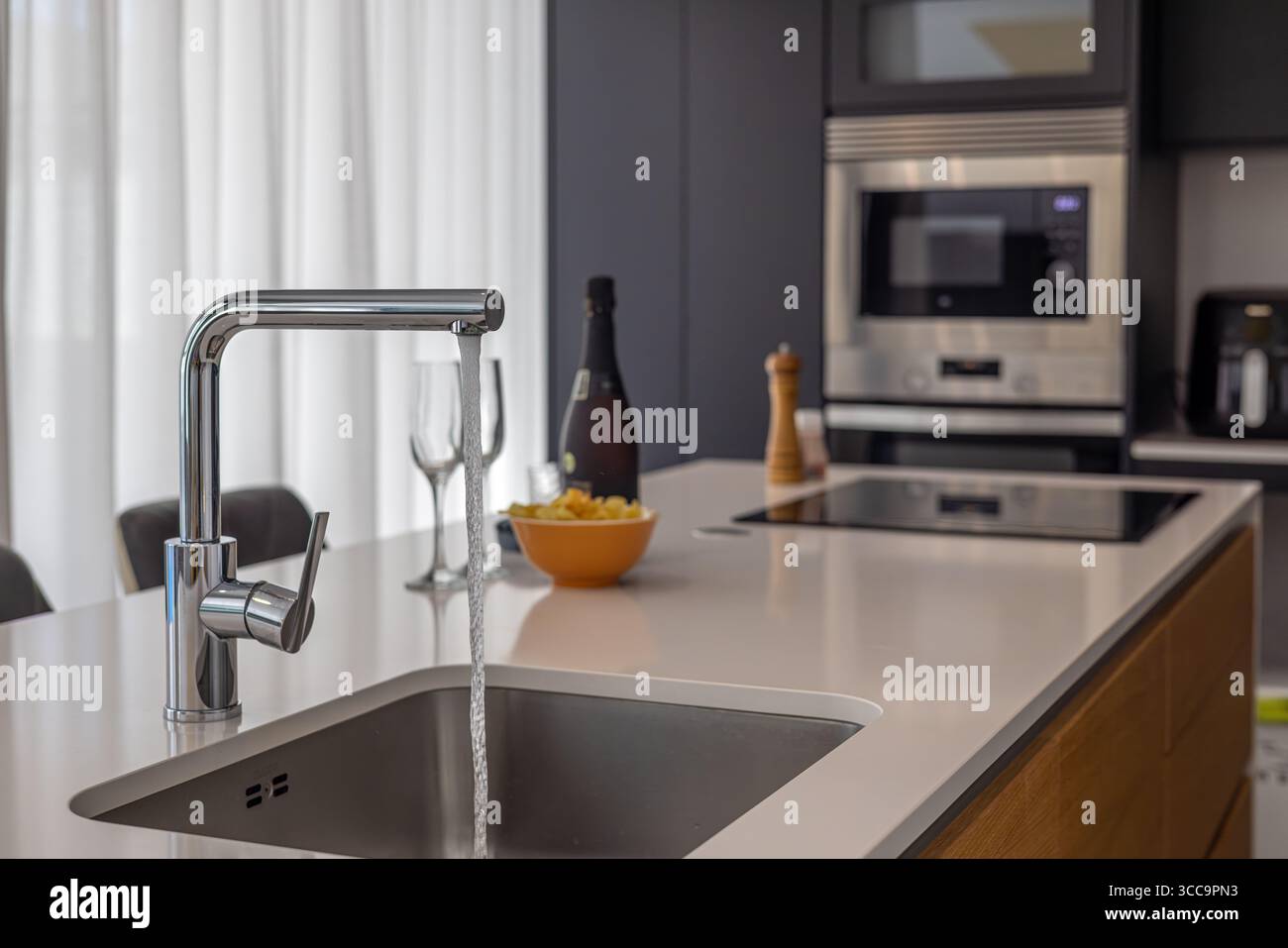 Cucina moderna a isola con rubinetto cromato con acqua corrente, piano cottura a induzione e accessori eleganti in uno spazio ben illuminato. Foto Stock