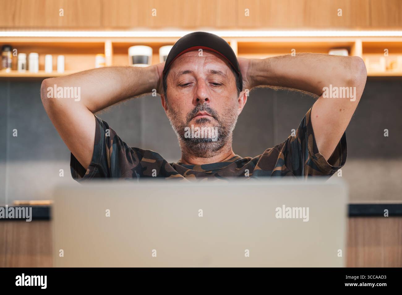 Uomo di mezza età con camicia mimetica e berretto seduto a casa con le mani dietro la testa, guardando lo schermo del notebook con un'espressione seria Foto Stock