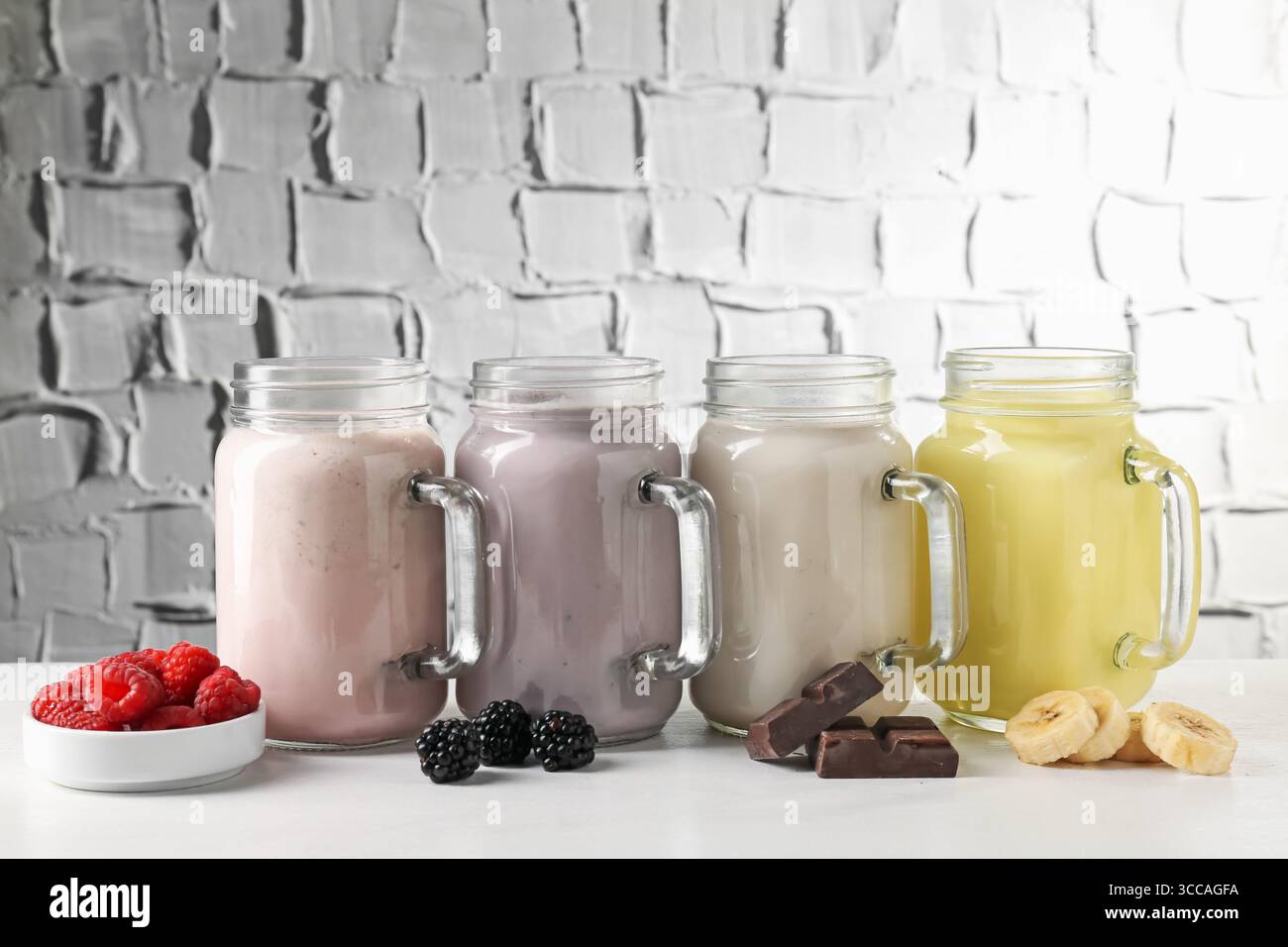 Diversi frullati gustosi nei vasetti mason e ingredienti sul tavolo bianco Foto Stock