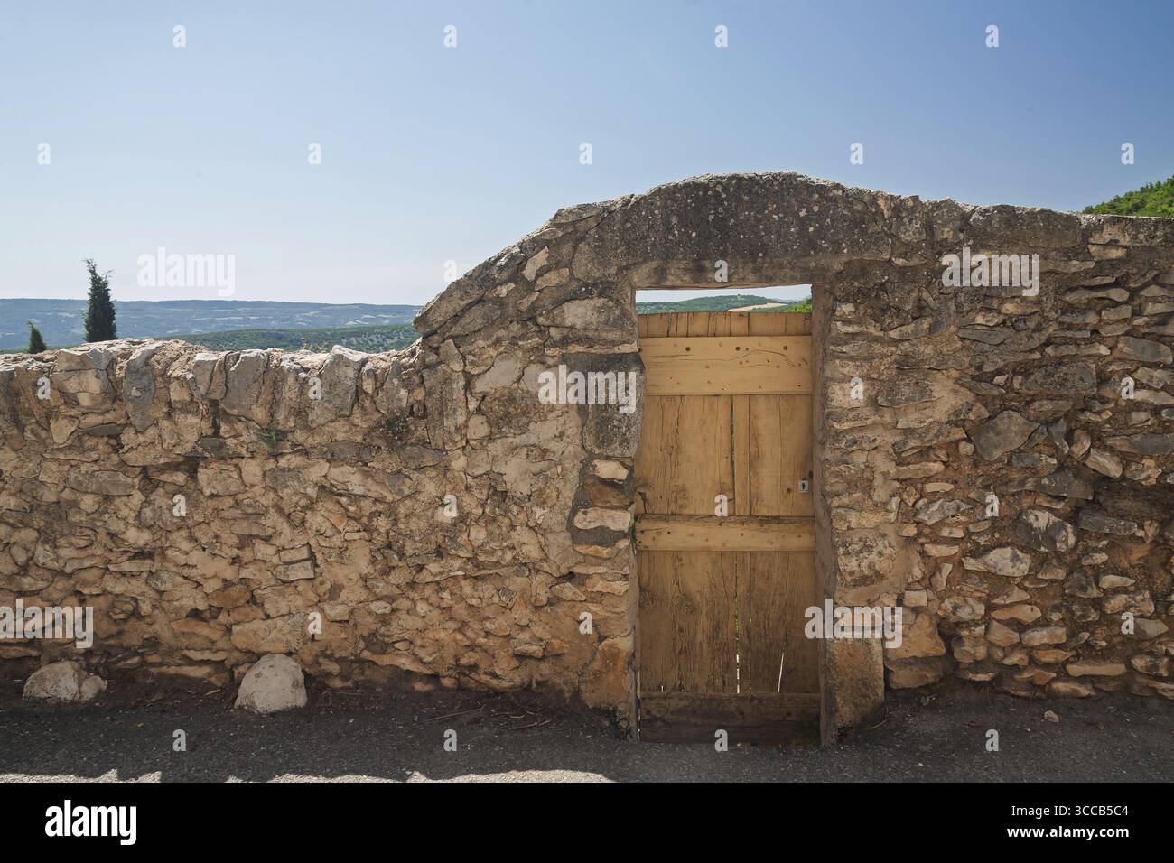 Muro in pietra con cancello in legno e vista di un ampio paesaggio sotto un cielo blu, Provenza, Francia Foto Stock